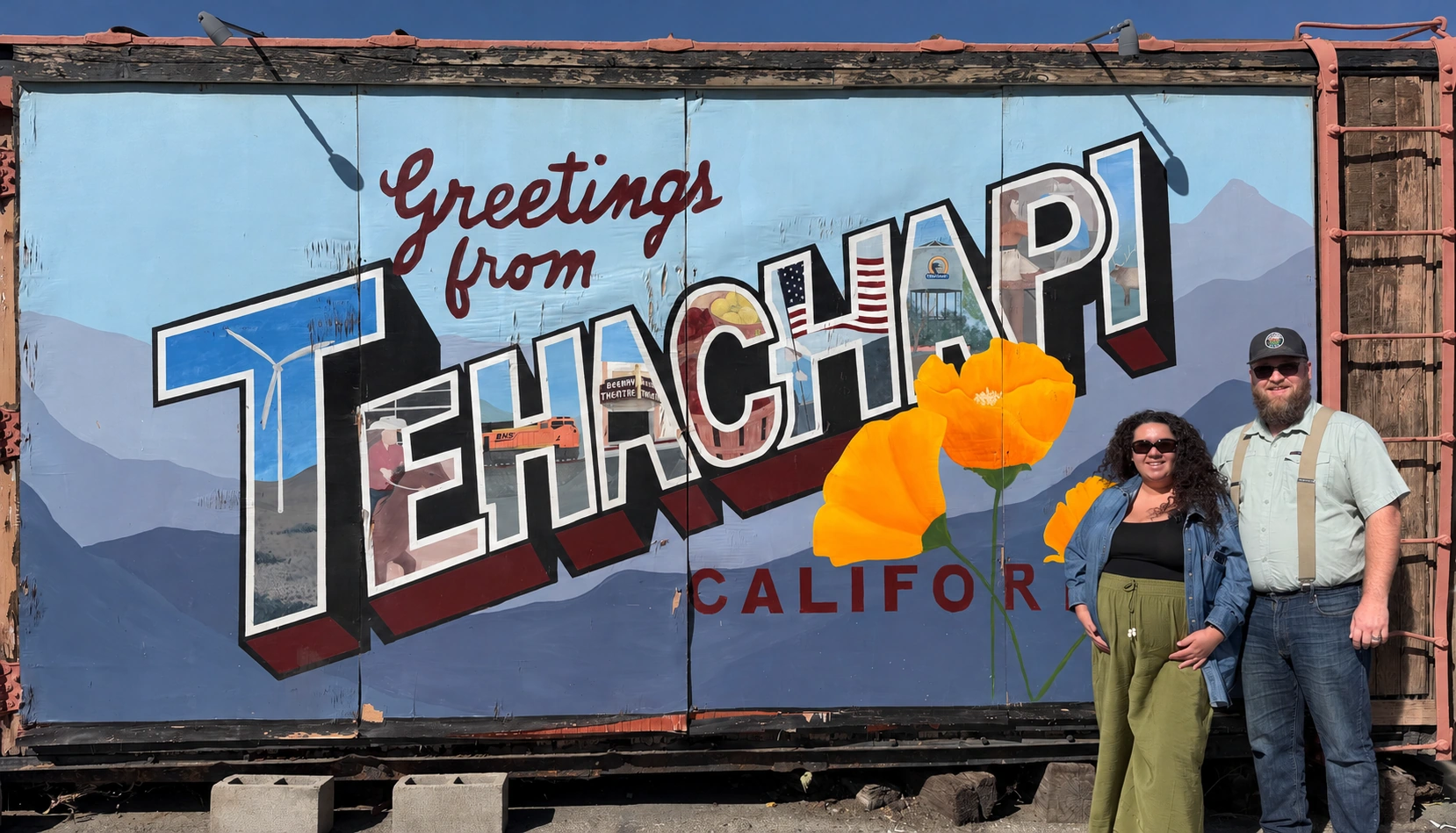 Laura and Bryce standing together beside the bright California poppies on the Greetings from Tehachapi railcar mural.